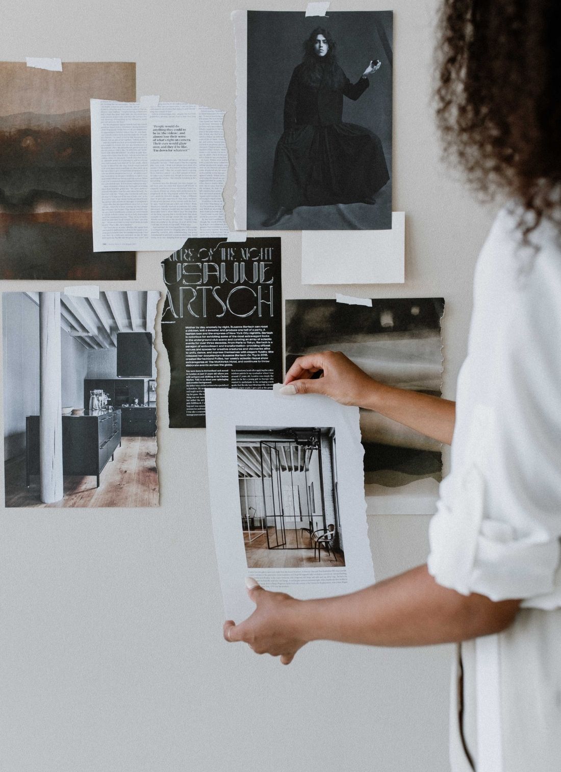 A person arranges a collection of monochrome and grayscale photographs and articles on a wall, showcasing artistic and architectural themes, illustrating the use of color psychology in branding.