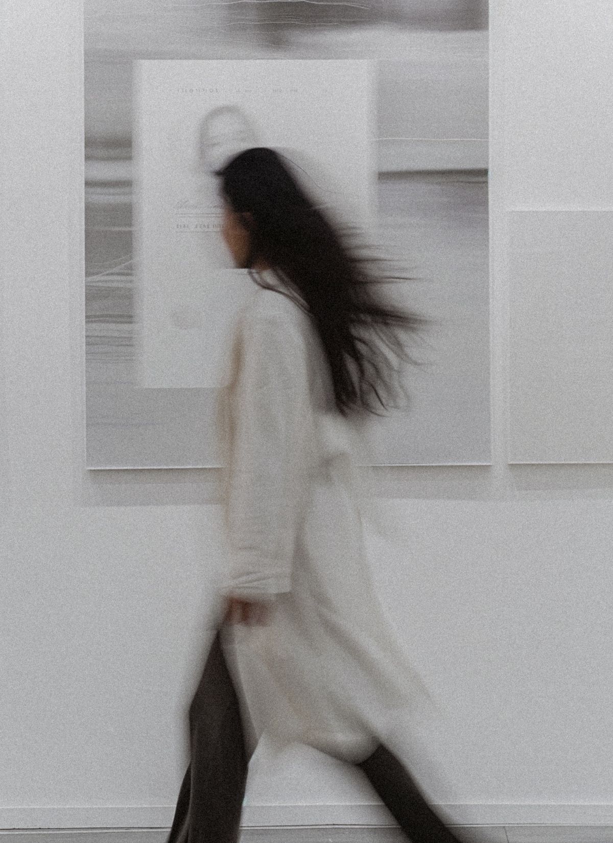 Woman in long coat walking through an art gallery, captured in soft motion blur with neutral tones and abstract art on the walls