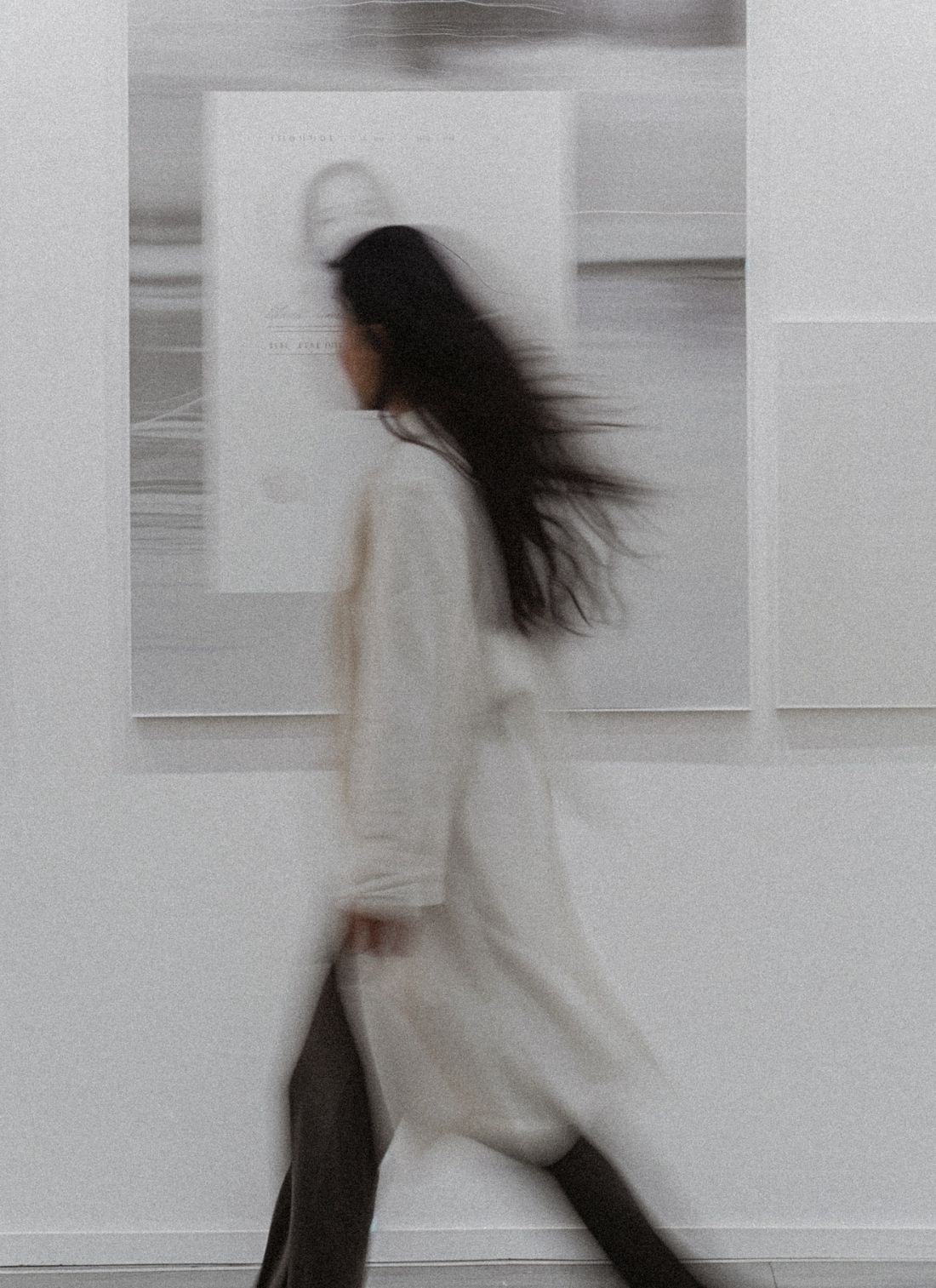 Woman in long coat walking through an art gallery, captured in soft motion blur with neutral tones and abstract art on the walls